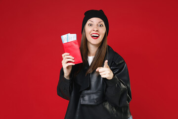 Wall Mural - Excited traveler tourist woman in leather jacket hat hold passport tickets point index finger on camera isolated on red background. Passenger traveling abroad on weekends. Air flight journey concept.