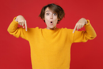 Wall Mural - Amazed surprised shocked young brunette woman 20s wearing casual yellow sweater standing pointing index fingers down on mock up copy space isolated on bright red colour background studio portrait.