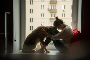 Wall Mural - A young woman with her shaggy dog at home, sits on a windowsill overlooking the city. Love for pets, joy and tenderness