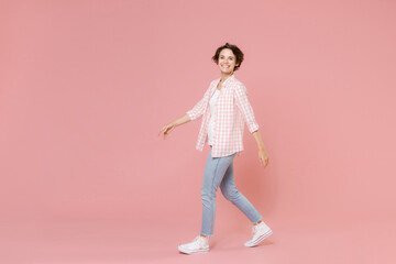 Full length side view of smiling beautiful attractive young woman 20s wearing casual basic checkered shirt walking going looking camera isolated on pastel pink colour background, studio portrait.