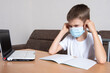 © Alena - A child in a protective mask on his face looks displeasedly at a laptop, a boy learns remotely online at home