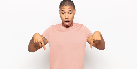 Wall Mural - young black man feeling shocked, open-mouthed and amazed, looking and pointing downwards in disbelief and surprise