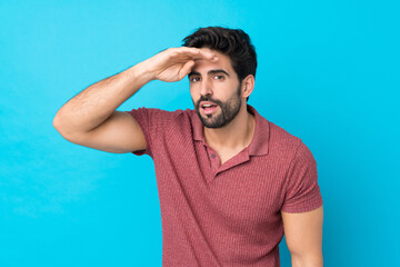 Wall Mural - Young handsome man with beard over isolated blue background looking far away with hand to look something