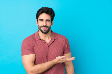 Wall Mural - Young handsome man with beard over isolated blue background presenting an idea while looking smiling towards