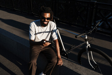 Wall Mural - African american guy smiling and using cellphone while sitting at street