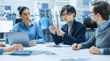 Factory Office Meeting Room: Computer Design Specialist Holds Printed Circuit Board Prototype and Reports to Diverse Team of Engineers, Managers Sitting at Conference Table. Industrial Facility