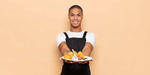 Sticker - young black man smiling happily with friendly, confident, positive look, offering and showing an object or concept