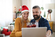 © Halfpoint - Happy couple with wine indoors at home at Christmas, having video call with family.