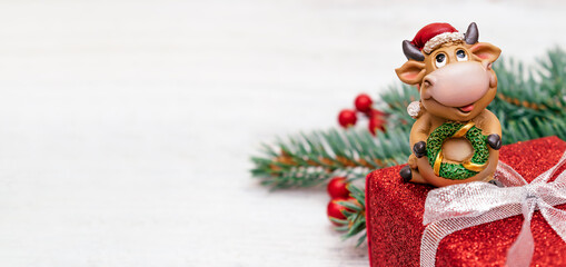 Toy bull in a Santa Claus hat on a red gift box on a white wooden background.Christmas tree branch with red berries in the background.
Bull as a symbol of the New year 2021.New Year,Christmas concept