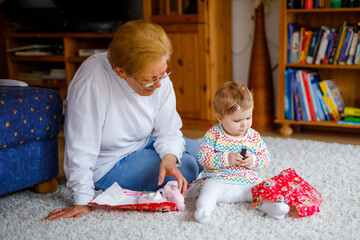 Wall Mural - Cute little toddler girl and grandmother playing with toys at home. Adorable baby child and senior retired woman unpacking gifts for birthday. Happy family together