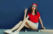 © rohappy - Attractive woman sitting on skateboard showing peace sign gesture wearing baseball cap over blue background