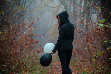 Wall Mural - Sad girl walking in dark clothing in autumn forest, with balloons, black and white. Concept of choice, good and evil