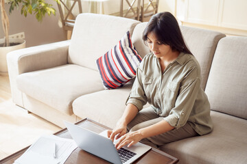 Young adult indian woman, female college student, remote social distance worker using laptop computer working online at home office, typing, e learning in internet sitting on couch in living room.