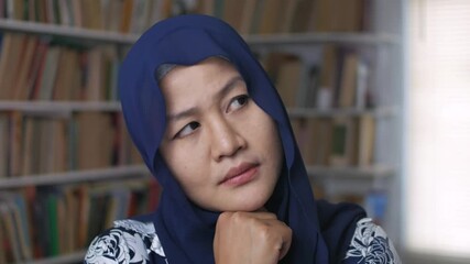 Wall Mural - Asian muslim woman thinking and smiling while standing in front of bookshelf, education, learning studying in library