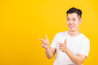 © sorapop - Portrait happy Asian handsome young man smiling standing wearing white t-shirt pointing finger to the side away he looking to space, studio shot isolated yellow background