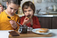 Sister And Brother Alone In The Kitchen Have Breakfast On Their Own Chocolate Paste, Smeared On The Toast Of Bread At The Table In The Kitchen. Children Have Fun Eating Chocolate Mousse