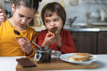 Sister and brother alone in the kitchen have breakfast on their own chocolate paste, smeared on the toast of bread at the table in the kitchen. Children have fun eating chocolate mousse