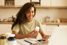 Biohacking And Superfoods Concept. Cheerful Young Latin Female Nutritionist Holding Bottle Of Dietary Supplements For Muscle Building. Pretty Girl Using Laptop Ordering Vitamins Via Online Store