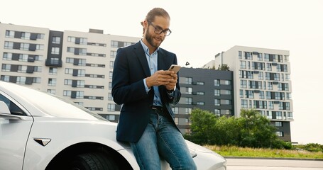 Happy Caucasian handsome male in glasses standing near modern electric car and texting on smartphone outddors. Young man in good mood browsing on cellphone. Urban concept