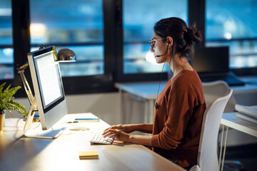 Beautiful young business woman working with computer while talking with earphone sitting in the office.