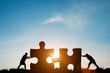 © Dilok - Silhouette two men pushing to connect jigsaw puzzle with sunlight and blue sky. It is symbol of business corporate teamwork to  solutions, success and strategy concept.