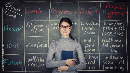Young woman teacher or student, wearing eyeglasses and holding a book, stands in front of blackboard written with chalk english grammar table with the list of tenses. Learning verb structure system.