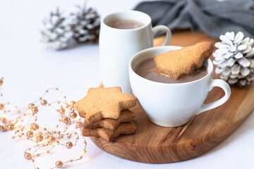 Two white cups with hot chocolate or cocoa drink with Christmas tree shape cookie on a wooden board and light marble background with festive winter decorations. Winter mood. Close up. Selective focus 