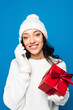 © LIGHTFIELD STUDIOS - pleased woman in knitted hat and gloves holding gift box and talking on smartphone isolated on blue