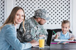 © LIGHTFIELD STUDIOS - Smiling wife looking at camera, typing on laptop, while sitting at table with blurred military man and girl on background