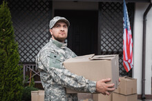 Military Serviceman Holding Cardboard Box, While Looking Away Near House And American Flag