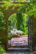 Arched Wrought Iron Gate in a Brick Wall Draped with Vines Peering into an inviting Verdant Garden with Brick Path 