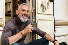 Smiling Happy Senior Male Traveler With Beard And Tattoo Drinking Coffee In The Doorway Of His Caravan