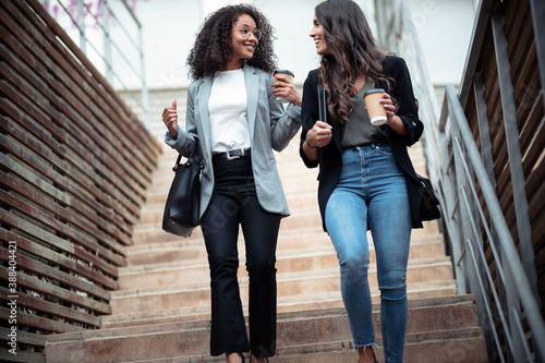 Two beautiful businesswomen talking and drinking coffee while going down the stairs in the street.