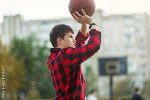 Cute young boy plays basketball on street playground. Teenager in red check flannel shirt with orange basketball ball outside. Hobby, active lifestyle, sport activity for kids.