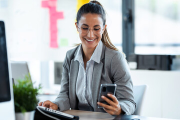 Wall Mural - Beautiful young business woman using smart phone while working with her computer in the office.