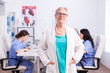 © DC Studio - Portrait of senior doctor smiling at camera in hospital conference room with medical staff in the background.