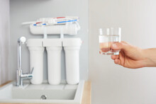 Men's Hand Holds A Glass Of Clear Water. Tap And Reverse Osmosis Filter In The Background.