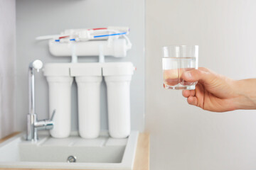 Men's hand holds a glass of clear water. Tap and reverse osmosis filter in the background.