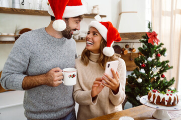 Wall Mural - Charming cheerful couple in Christmas hats smiling and using cellphone