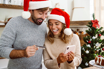 Wall Mural - Nice happy couple in Christmas hats using credit card and cellphone