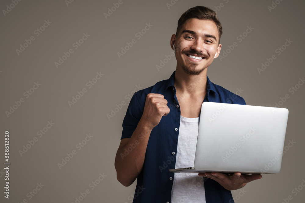 Image of happy handsome guy showing winner gesture while holding laptop - obrazy, fototapety, plakaty 