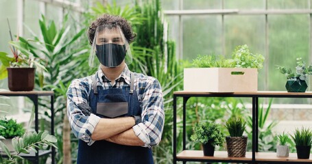 Wall Mural - Portrait of Caucasian handsome joyful man owner of flower shop in face shield and mask smiling to camera indoors. Cheerful young man worker standing in greenhouse in good mood. Employment concept