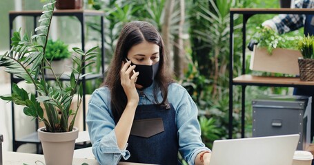 Wall Mural - Portrait of beautiful Caucasian woman worker in apron and mask talking smartphone and typing on laptop in flower shop. Female florist checking flowers indoors and calling on cellphone. Work concept