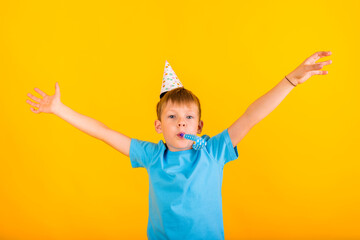 portrait of a boy toddler sitting in a paper holiday cap and whistle and spread his hands to the sides on a yellow background