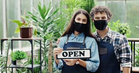 Wall Mural - Close up portrait of happy young Caucasian male and female florists holding Open sign with smile on faces. Joyful flower shop employees in masks posing to camera indoors. Floral business concept