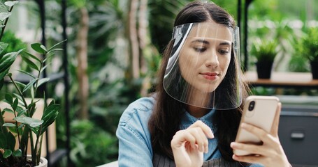 Wall Mural - Close up portrait of beautiful Caucasian female businesswoman in face shield sitting in flower shop and tapping on mobile phone. Young woman worker of floral center texting on smartphone. Job concept