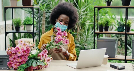 Wall Mural - Beautiful African American female businesswoman in face mask sitting in flower shop and tapping on computer while checking flower. Woman employee of garden center browsing on laptop. Floral concept