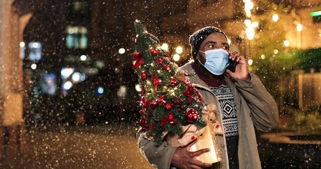 Close up portrait of joyful man standing on snowy street and calling on cellphone. Handsome African American male speaking on smartphone outdoor and holding little new year tree. Holidays concept