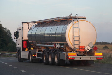 Gray semi truck tanker with 30 1866 dangerous class sign on barrel drive on asphalt highway on a summer day on green trees background, liquid tar ADR hazardous cargo logistics, rear view