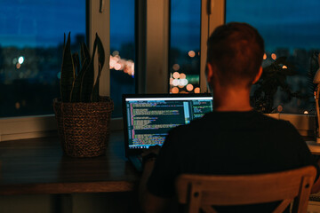 Wall Mural - a young male programmer in front of a window with a lights of night city, on his laptop screen is co
